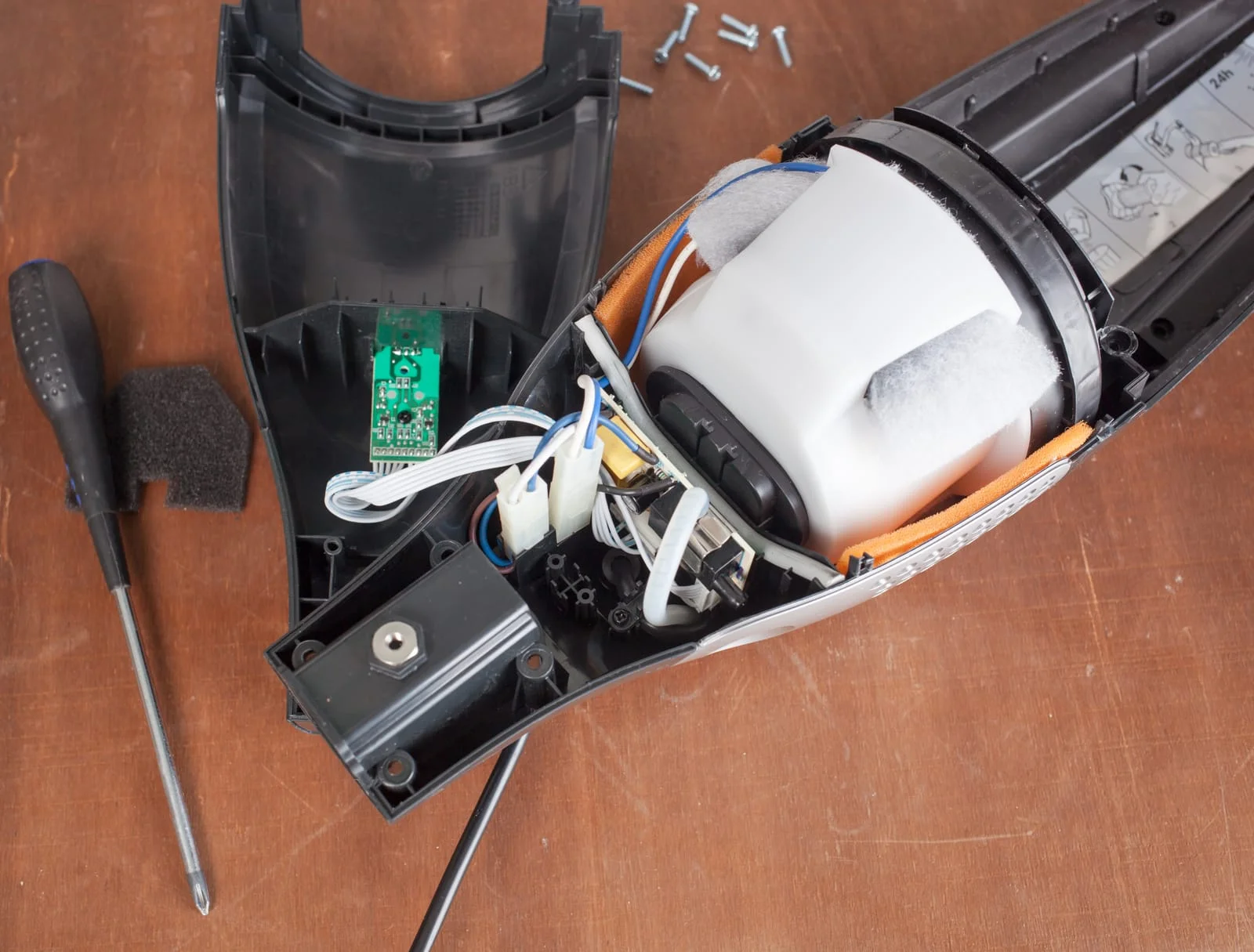 Open handheld device showing its internal components: circuit board, ribbon cables, foam padding, and screws on a wooden table with a screwdriver nearby.